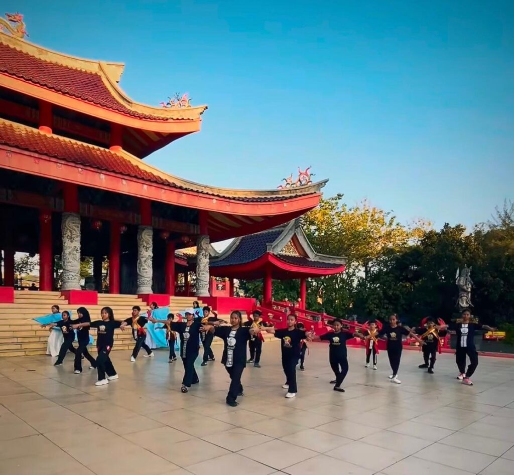 Whatsapp image 20260616 at 15 08 12 catrawarta Group of people dancing in black outfits on a tiled courtyard in front of a red asian temple with curved roofs under a blue sky